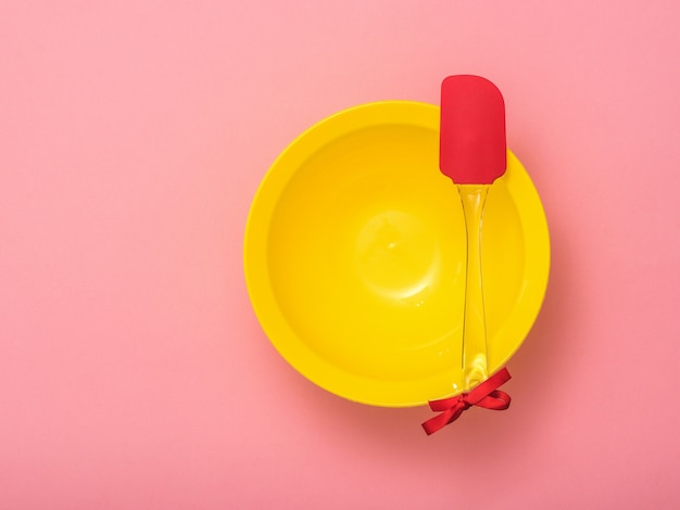 Spatola da cucina con nastro rosso e ciotola gialla su sfondo rosa. Utensili da cucina su sfondo festivo. Disposizione piatta.