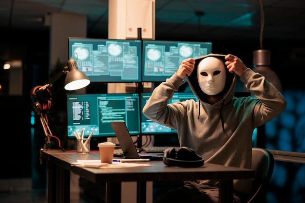 Spaventoso hacker mascherato che installa virus per hackerare il sistema sul computer, lavorando a tarda notte in ufficio. Cyber terrorista e criminale con maschera che hackera il server di rete e fa truffe o frodi.