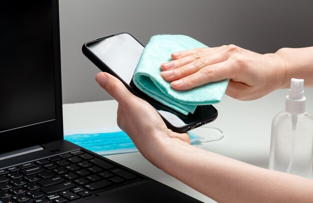 Spazio di lavoro dello smartphone pulito della donna. Disinfezione della tastiera del telefono e del laptop con disinfettante alcolico. Superfici della scrivania dell'ufficio del posto di lavoro di pulizia della donna Nuova igiene normale covid 19 coronavirus.