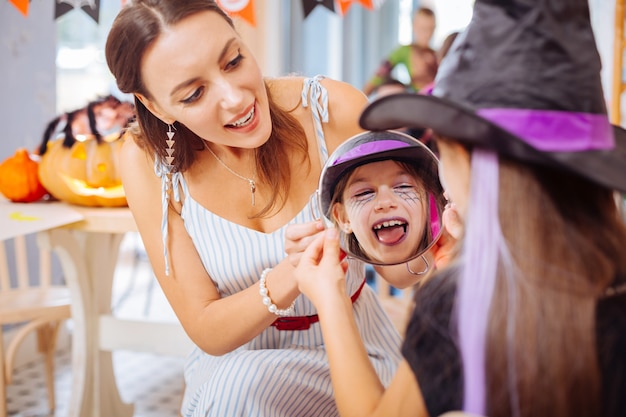 Specchio e trucco. Bella madre alla moda amorevole che tiene miracolo che mostra alla figlia il suo trucco di Halloween