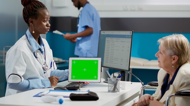 Specialista che mostra lo schermo verde orizzontale sul tablet a una donna anziana paralizzata, guardando il modello chroma key isolato sul display. Medico e paziente in sedia a rotelle utilizzando copyspace mockup vuoto.