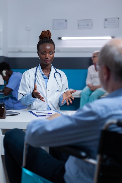 Specialista in clinica che discute con un pensionato in sedia a rotelle sugli effetti dell'antivirale prescritto. Medico che spiega i benefici dei farmaci e i risultati del recupero alla persona anziana