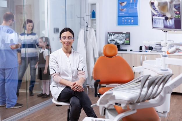 Specialista sanitario in igiene dei denti che indossa camice bianco seduto su uno sgabello e assistente che discute con i pazienti. Stomatolog nella clinica dei denti professioanl sorridente indossando l'uniforme che guarda l'obbiettivo.