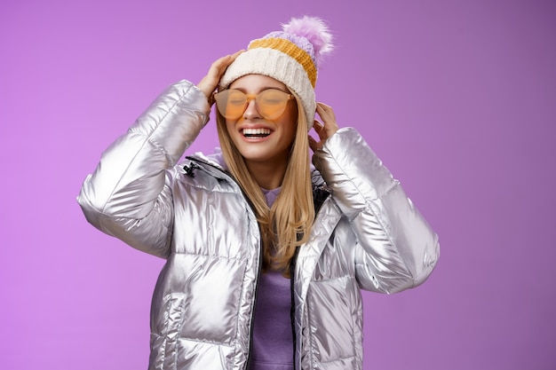 Spensierata e divertita affascinante ragazza bionda divertendosi godendo di una fantastica giornata invernale di sole vacanza in località sciistica indossando occhiali da sole giacca elegante argento messo sul cappello sorridendo con gioia, sfondo viola