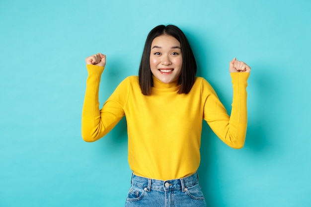 Speranzosa ragazza asiatica che vince il premio, stringe i pugni e sorride felice alla telecamera, trionfando del successo e del successo, in piedi su sfondo blu.