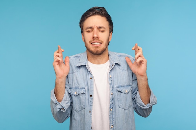 Spero, vinco io. Ritratto di uomo in camicia di jeans da lavoratore incrociando le dita e augurando buona fortuna, pregando prima della lotteria, in previsione del successo. tiro in studio indoor isolato su sfondo blu