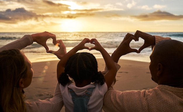 Spiaggia cuore mani e famiglia per amore dell'oceano estivo e benessere all'aperto con genitori bambino e nuvole del cielo al tramonto Dietro di persone o madre e padre con bambino con segno di cura o icona per il mare