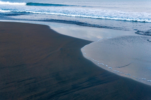 Spiaggia dell'oceano con sabbia vulcanica nera, il cielo si riflette nell'onda rovesciata del surf