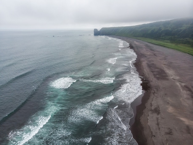 Spiaggia di sabbia di colore scuro quasi nero dell'oceano Pacifico. Le montagne di pietra e l'erba gialla sono su uno sfondo. Cielo azzurro.
