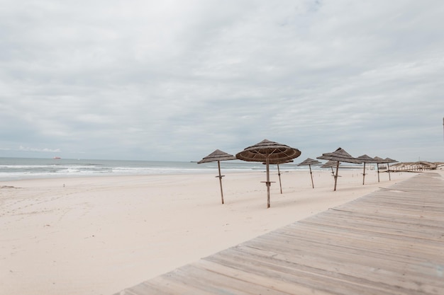 Spiaggia sabbiosa vuota con una passerella in legno e ombrelloni di paglia vicino all'oceano Vacanze in Portogallo Oceano Atlantico Vacanze al mare