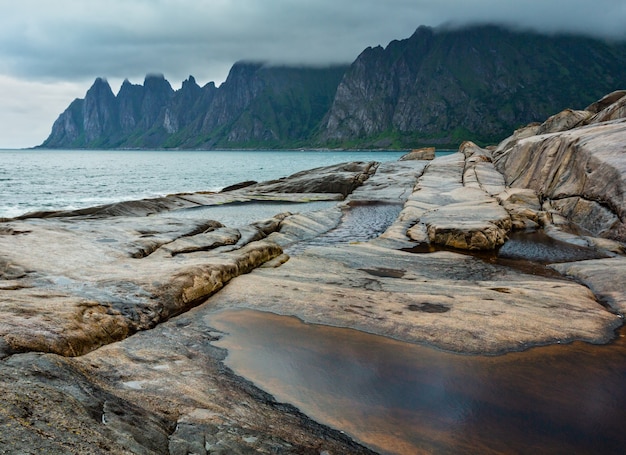 Spiaggia sassosa con bagni di marea a Ersfjord, Senja, Norvegia. Costa polare di notte di giorno di estate. I denti di drago oscillano lontano.