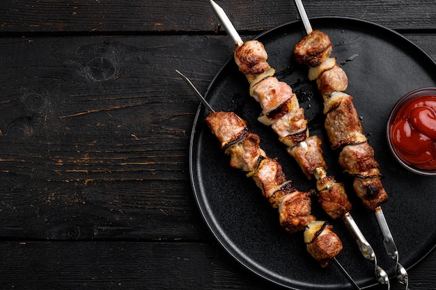 Spiedini succosi di carne di maiale o set di shashlik, su sfondo di tavolo in legno nero, vista dall'alto piatta, con spazio di copia per il testo