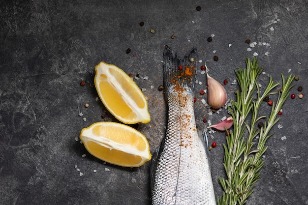 Spigola di pesce fresco e ingredienti per cucinare, limone e rosmarino. Vista dall'alto di sfondo scuro.