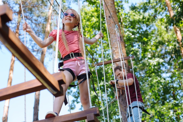 Spirito avventuroso. Piacevole bambina che cammina lungo un sentiero in un parco avventura e sorride fiduciosa mentre suo padre la segue