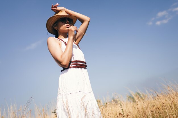 Spirito di libertà Un'attraente ragazza boho in camicetta cappello e occhiali da sole in piedi sul campo sullo sfondo di un cielo blu Vacanze estive in viaggio stile hippie moderno bohémien