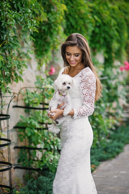 Splendida bella ragazza sorridente in elegante abito bianco di pizzo con un cagnolino bianco nelle mani