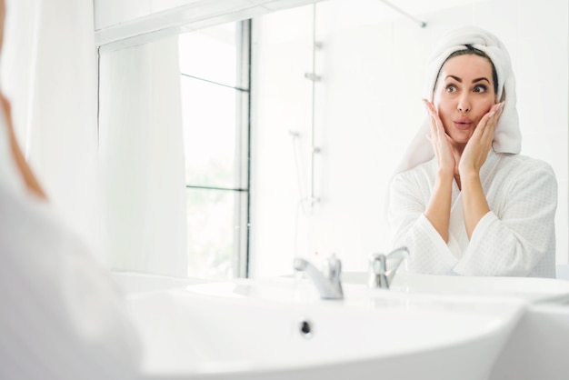 Splendida donna adulta sicura di sé che indossa un asciugamano sulla testa seduta e controllando la mascella davanti allo specchio nel bagno di casa Uno stile di vita di bellezza donna piuttosto attraente e concetto cosmetico