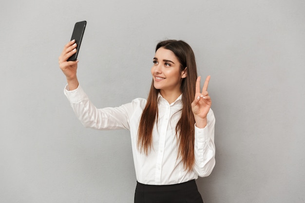 Splendida donna castana in abbigliamento business prendendo selfie sul cellulare e gesticolando segno di vittoria, isolato sopra il muro grigio