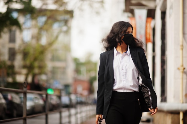 Splendida donna indiana indossa una maschera facciale formale e nera che posa in strada durante la pandemia covid con il cellulare a portata di mano