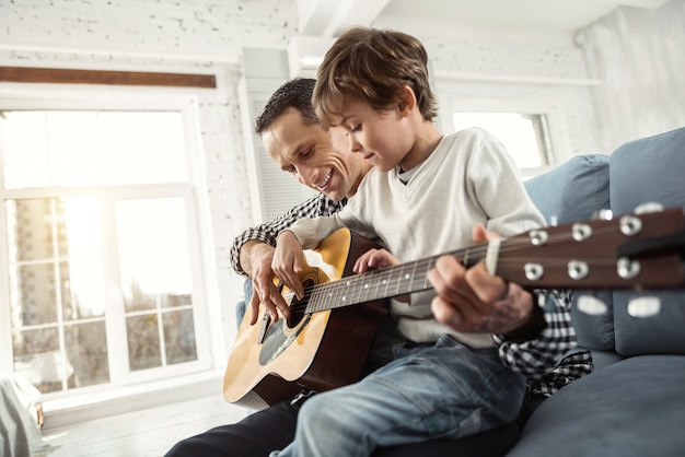 Splendida giornata. Ragazzo biondo contenuto attraente che impara a suonare la chitarra mentre era seduto sul divano e suo padre sorrideva