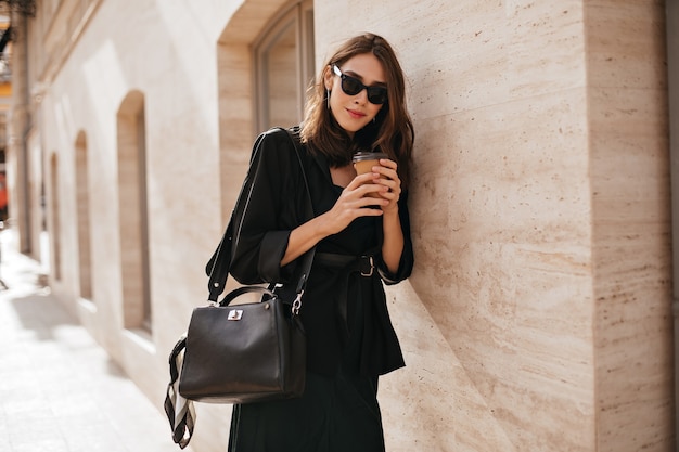 Splendida giovane donna con acconciatura ondulata bruna, occhiali da sole, cappotto nero e borsa che cammina nella città diurna e posa contro il muro beige