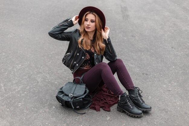 Splendida giovane donna dai capelli rossi con un cappello viola in una giacca di pelle in pantaloni alla moda con uno zaino alla moda