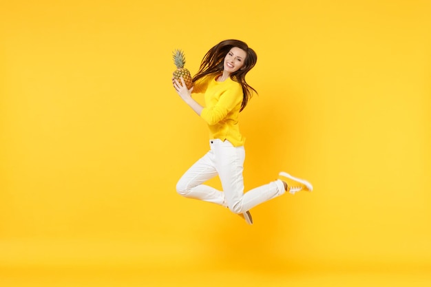 Splendida giovane donna in abiti casual che saltano, tengono frutta fresca di ananas matura isolata su sfondo giallo arancione parete in studio. Persone stile di vita vivido, relax concetto di vacanza. Mock up copia spazio