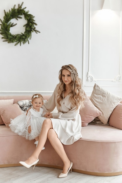 Splendida giovane donna in abito elegante in posa con la sua piccola figlia in abito soffice in interni decorati per Natale.