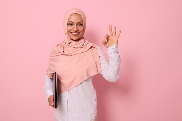 Splendida giovane donna musulmana di etnia mediorientale che tiene un computer portatile sotto le braccia e mostra il segno OK con il dito, sorride con un sorriso a trentadue denti che guarda l'obbiettivo, sfondo rosa copia spazio