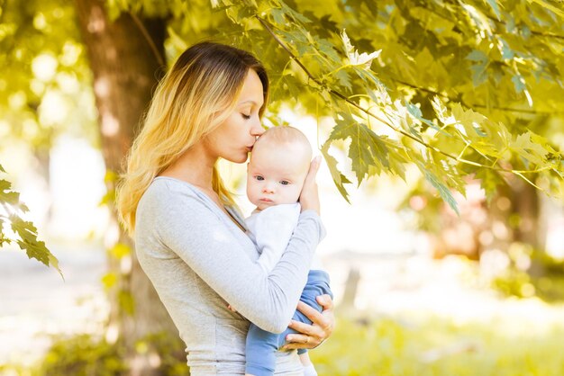 Splendida madre premurosa che tiene bambino neonato, baciandogli la testa. Madre calma e piena di amore che bacia la testa del suo bambino, in piedi all'aperto nel parco estivo. Bambino che guarda la telecamera