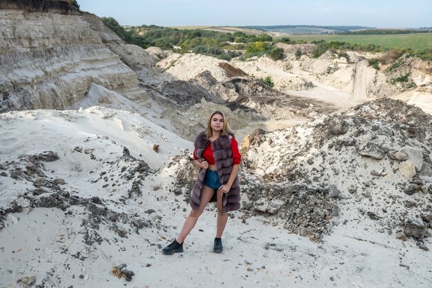 Splendida moda modello indossare cappotto di pelliccia in piedi alla cava di sabbia vuota secca nella calda giornata di sole autunnale