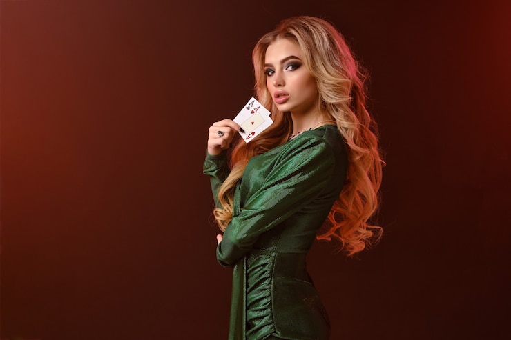 Foto splendida modella bionda, trucco luminoso, in abito elegante verde e gioielli. guardandoti, mostrando due carte da gioco, in posa di lato su sfondo marrone, retroilluminazione. gioco d'azzardo, poker, casinò. avvicinamento