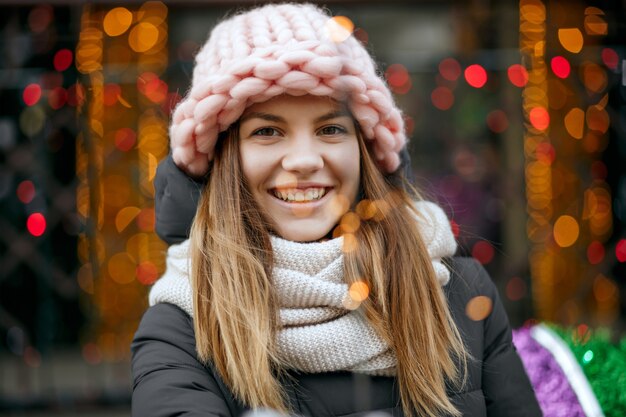 Splendida ragazza bionda sorridente che indossa abiti invernali che celebrano il Natale alle ghirlande sfondo sfocato. Spazio per il testo