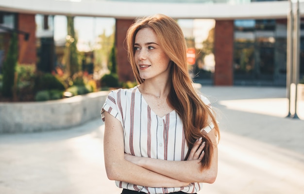 Splendida ragazza caucasica dai capelli rossi con le lentiggini in posa con le mani incrociate mentre distoglie lo sguardo