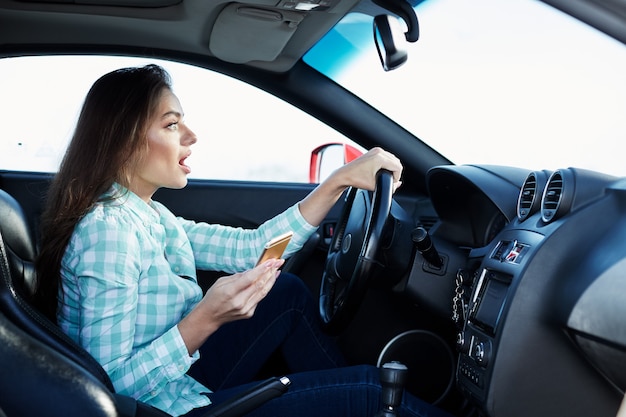 Splendida ragazza che indossa la camicia blu seduta nella nuova automobile, felice, bloccata nel traffico, ascoltando la musica, ritratto, tenendo il cellulare, incidente.