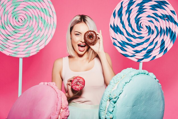 Splendida ragazza con capelli biondi che indossa top e berretto rosa in piedi con enormi dolci lecca-lecca in studio rosa