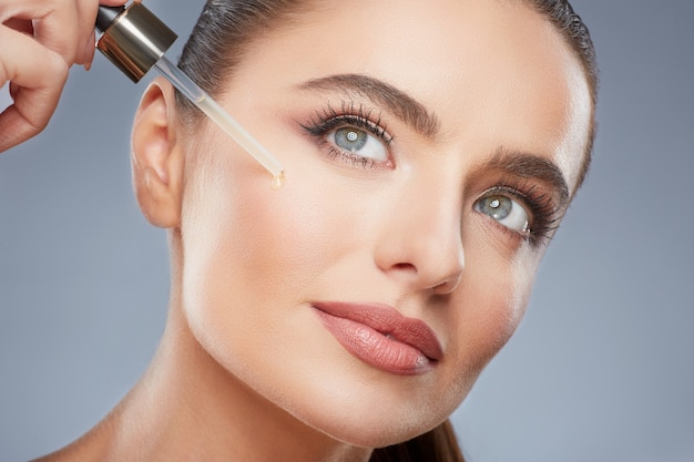 Splendida ragazza con i capelli castani fissati dietro, pelle pulita e fresca, grandi occhi e spalle nude che posano sullo sfondo grigio dello studio, foto di bellezza, tenendo l'olio per il viso, alzando lo sguardo.