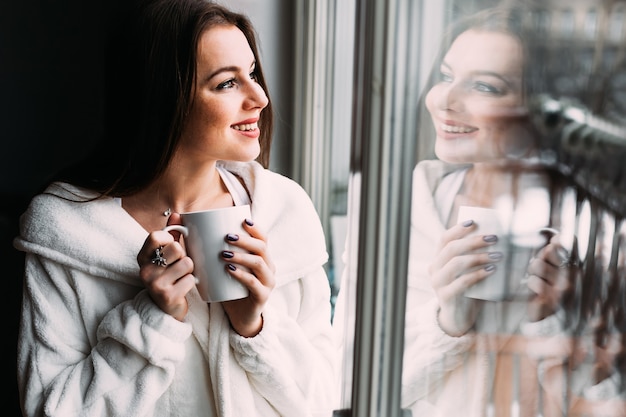 Splendida ragazza di sogno sorridente in vestaglia bianca stare vicino alla finestra con una tazza di caffè o tè. Caffè del mattino, concetto di felicità.