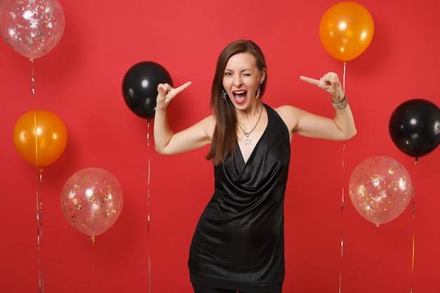 Splendida ragazza in abito nero che celebra il dito indice puntato lampeggiante su se stessa su mongolfiere di sfondo rosso brillante. San Valentino, concetto di festa di compleanno mockup di felice anno nuovo.