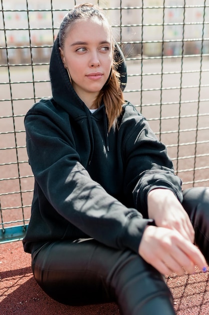 Splendida ragazza in pullover nero con cappuccio che guarda da parte la mano mentre riposa all'aperto Modello femminile dai capelli lunghi ispirato con grandi occhi verdi k seduto per terra