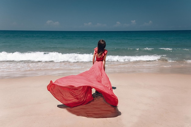 Splendida ragazza nell'esotica spiaggia sabbiosa in riva al mare. La signora indossa un abito rosso chiaro che scorre al vento; concetto di moda.