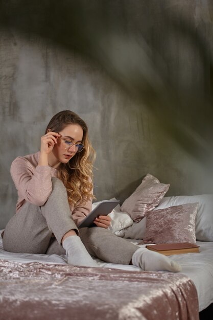 Splendida signora bionda con gli occhiali, abbigliamento casual. Seduto sul letto, lavorando con un tablet in camera da letto a casa. Studente, blogger, studio di documenti. Interno con parete grigia, coperta rosa e cuscini