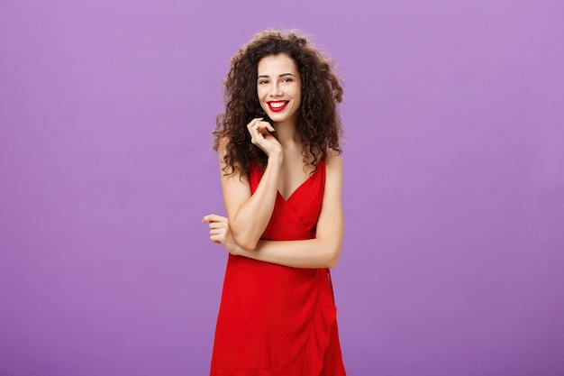 Splendida signora in rosso che si sente timida e imbarazzante essere invitata a ballare in piedi timida e femminile su sfondo viola toccando una ciocca di capelli ricci sorridendo ampiamente e incrociando il braccio sul petto.