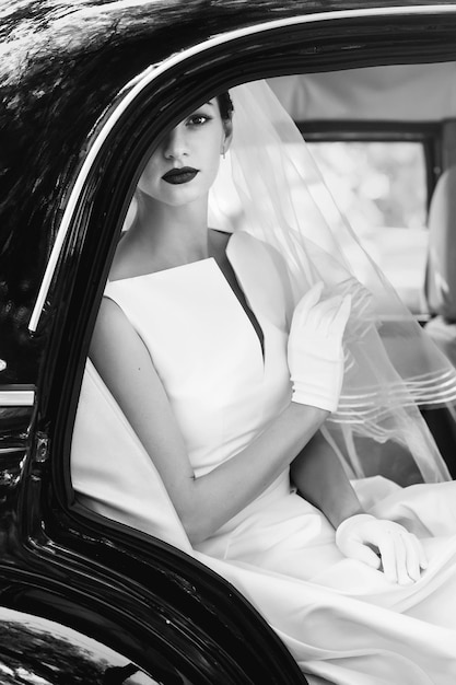 Splendida sposa elegante in posa in un'elegante macchina nera retrò seduta all'interno di un matrimonio di lusso nel salone in foto in bianco e nero con ritratto in stile vintage