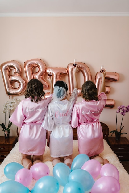 Splendide spose e damigelle brindano con il vino e si divertono la mattina del matrimonio Ragazze che tengono bicchieri con bevande e tintinnano su uno sfondo di palloncini Vista posteriore