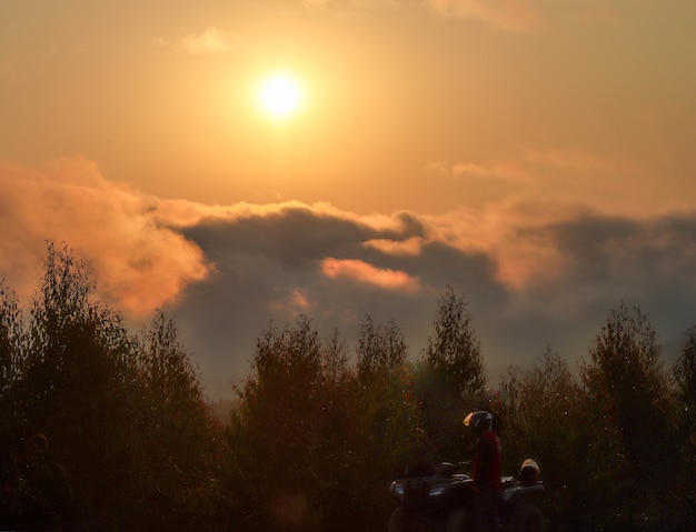 Splendido scenario di montagna e natura, tramonto sopra le nuvole, con il sole che splende attraverso le nuvole sullo sfondo e la sagoma di pini e vegetazione in primo piano.