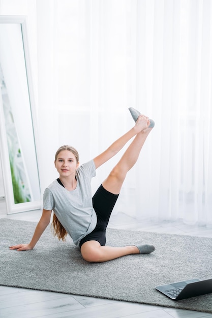 Sport a casa per bambini Ginnastica online Allenamento pilates virtuale Ragazza atletica che fa stretching esercizio fisico da solo sul pavimento con video tutorial sul laptop all'interno della stanza luminosa