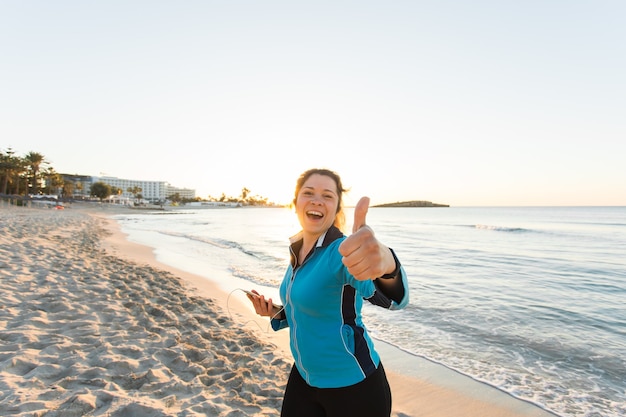 Sport all'aperto, gadget per il fitness e concetto di persone - Il fitness femminile sorridente mostra i pollici in su e tiene in mano lo smartphone con gli auricolari.