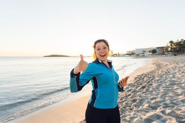 Sport all'aperto, gadget per il fitness e concetto di persone - Il fitness femminile sorridente mostra i pollici in su e tiene in mano lo smartphone con gli auricolari.