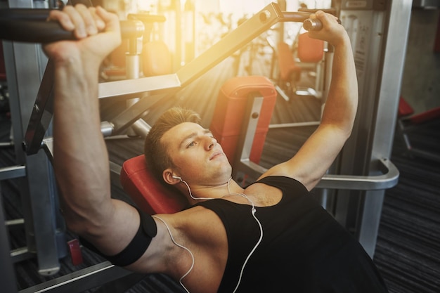 sport, attrezzatura, tecnologia, stile di vita e concetto di persone - giovane con auricolari che flettono i muscoli del torace sulla macchina da palestra